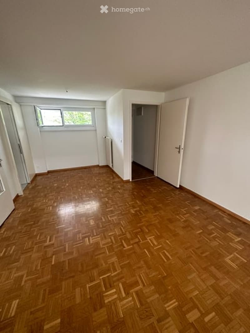 Empty room with wooden parquet flooring, white walls, and a single window letting in natural light. No furniture or equipment is present.