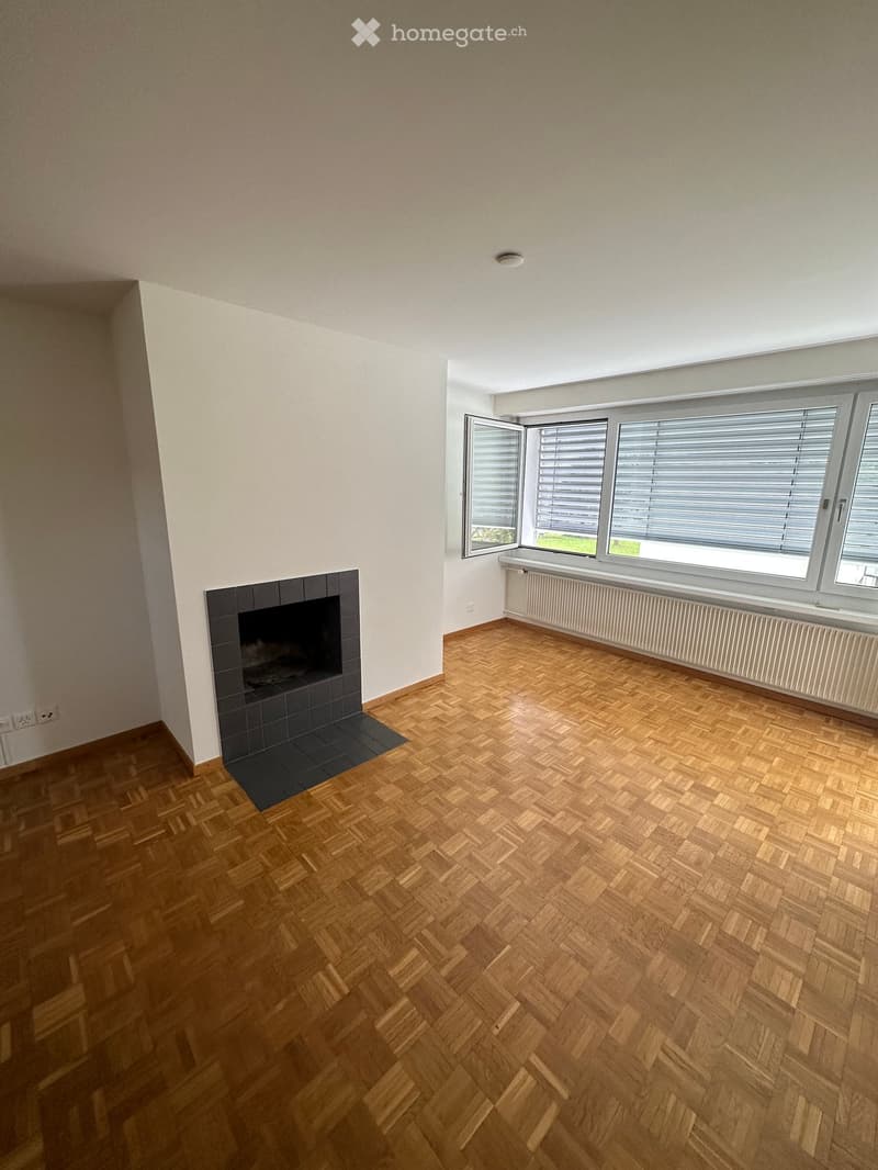 Empty room with wooden herringbone flooring, white walls, fireplace, and large windows with blinds.