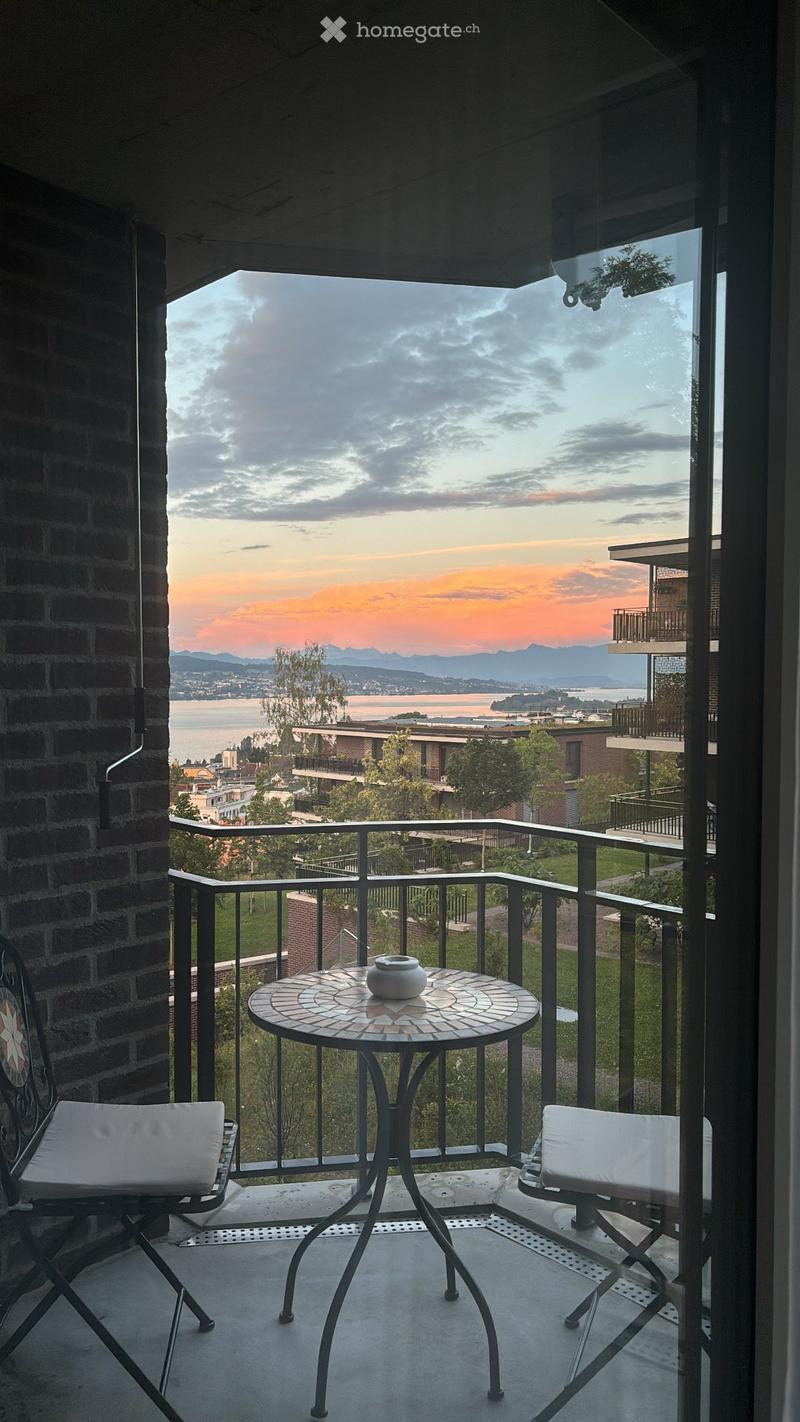Balcony with a round mosaic table, two white cushioned chairs, and a decorative vase, overlooking a scenic view of a lake and distant mountains.