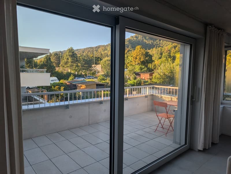 Balcony with tiled floor, metal railing, and a view of a forested area. A red folding chair and a small table are placed on the balcony.