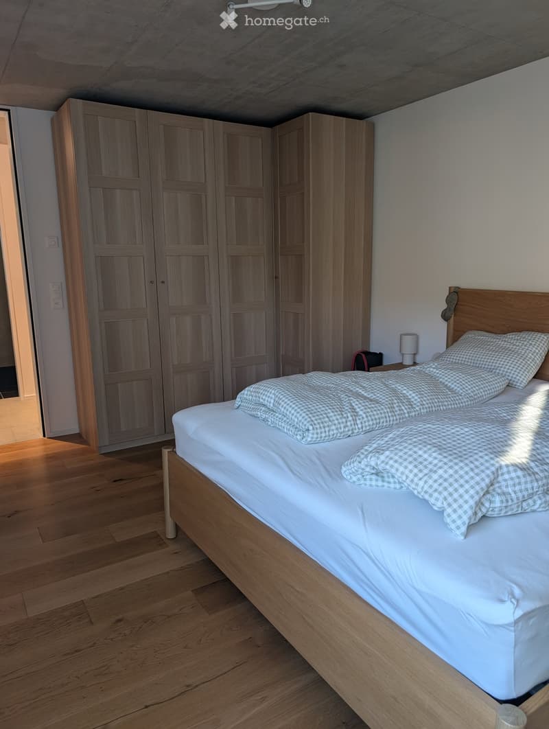 A well-lit bedroom with a wooden bed, white bedding, a large wooden wardrobe, and a nightstand with a lamp.