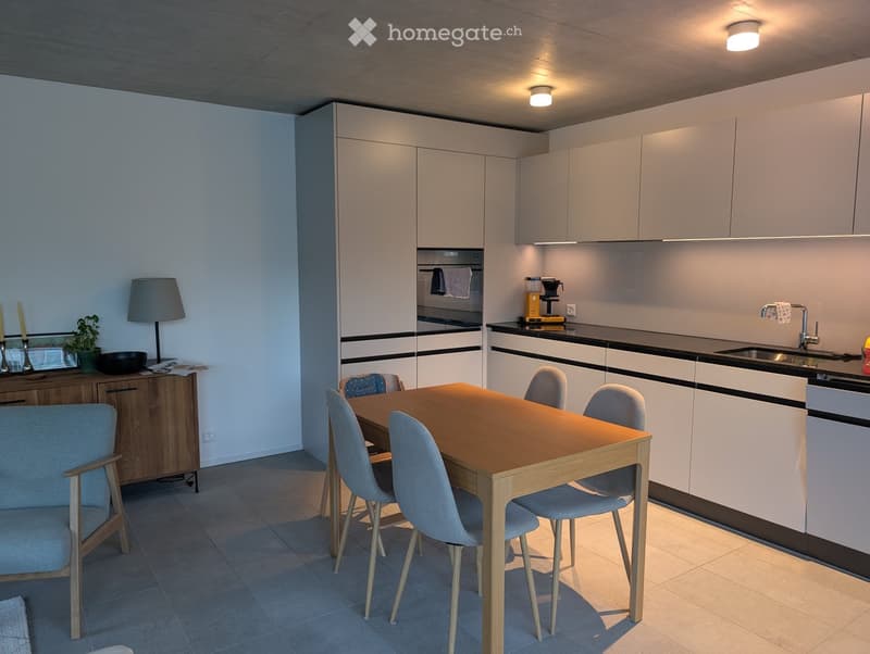 Modern kitchen and dining area with white cabinets, a wooden dining table, grey chairs, a sink, and a coffee maker on the counter.