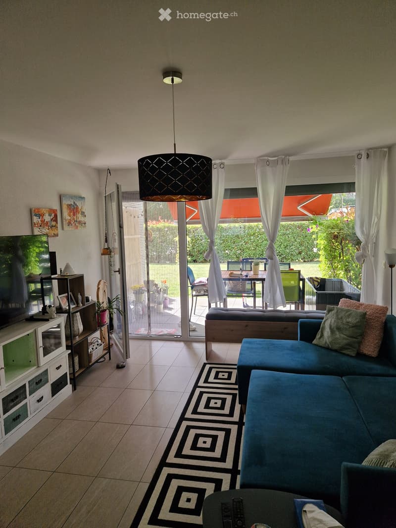 Modern living room with a blue sectional sofa, coffee table, TV on a shelf, pendant light, and an open sliding door leading to a patio with outdoor furniture.