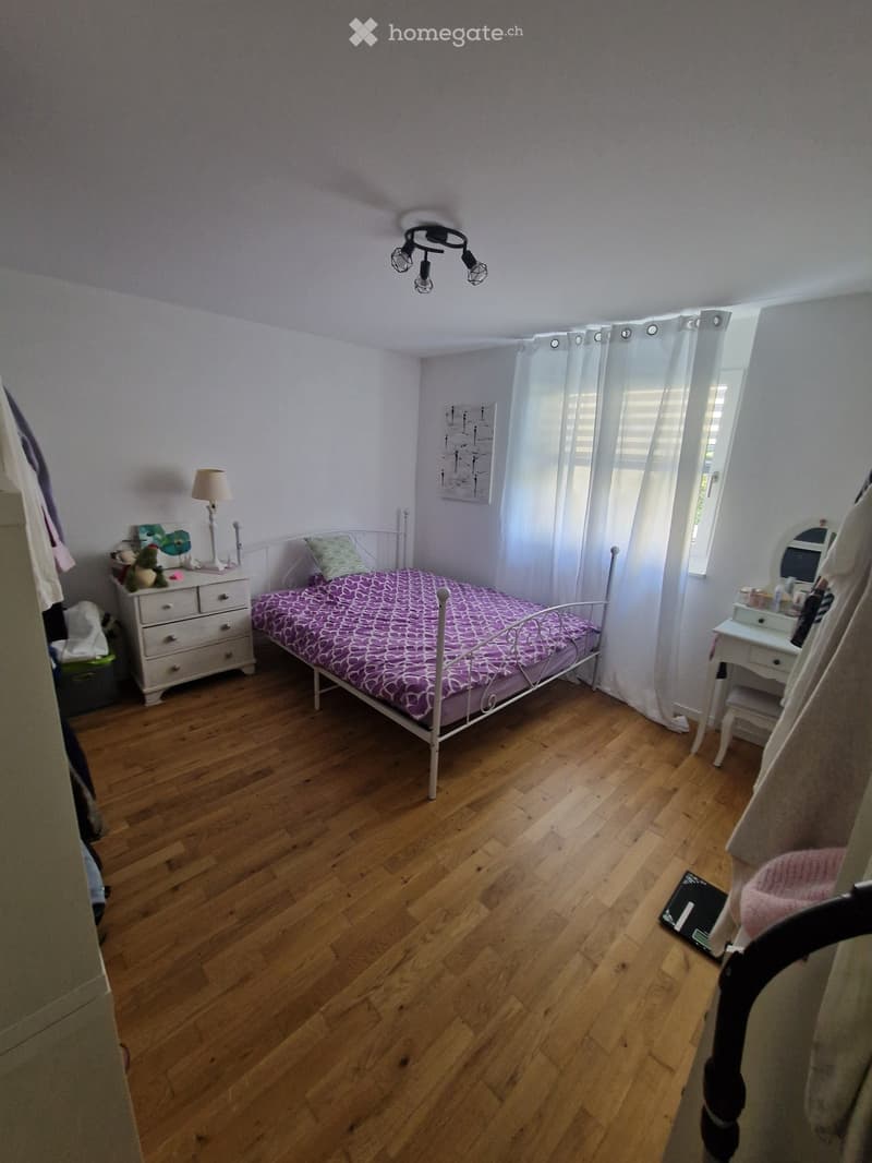 Cozy bedroom with wooden flooring, a single bed with a purple patterned bedspread, a nightstand with a lamp, a mirror on a small table, and a small stool near the window.
