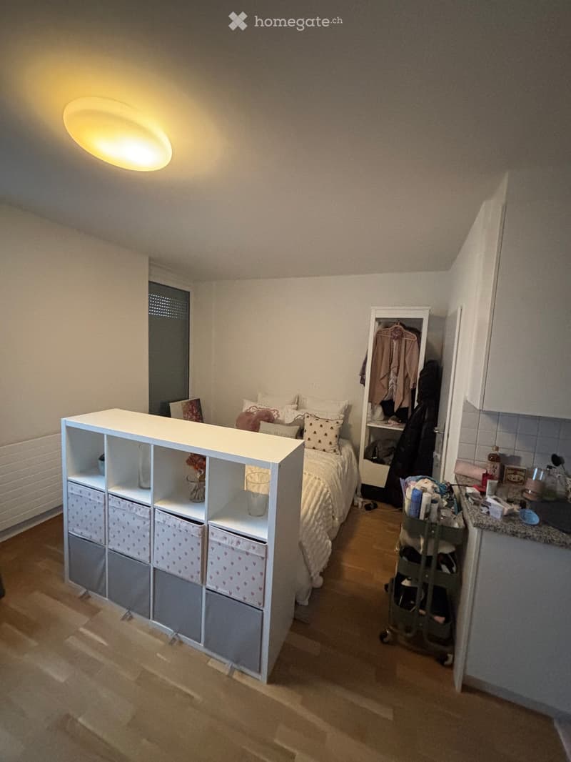 A small, cozy bedroom with a bed, storage unit, mirror, clothing rack, and a cart with shoes. The room features wooden flooring and a slanted ceiling with a round light fixture.
