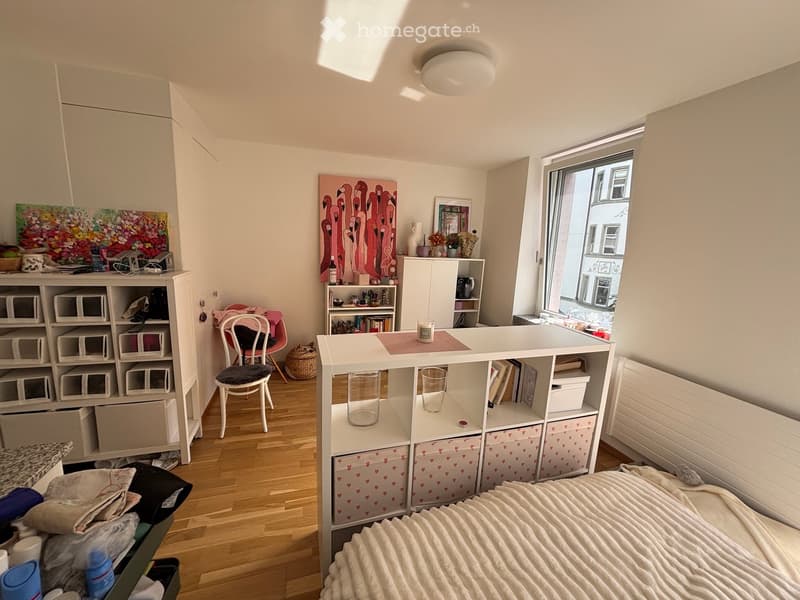 Cozy bedroom with a bed, a white table with two glasses, a chair, a shelf with storage boxes, and various decorative items including artworks and a vase.