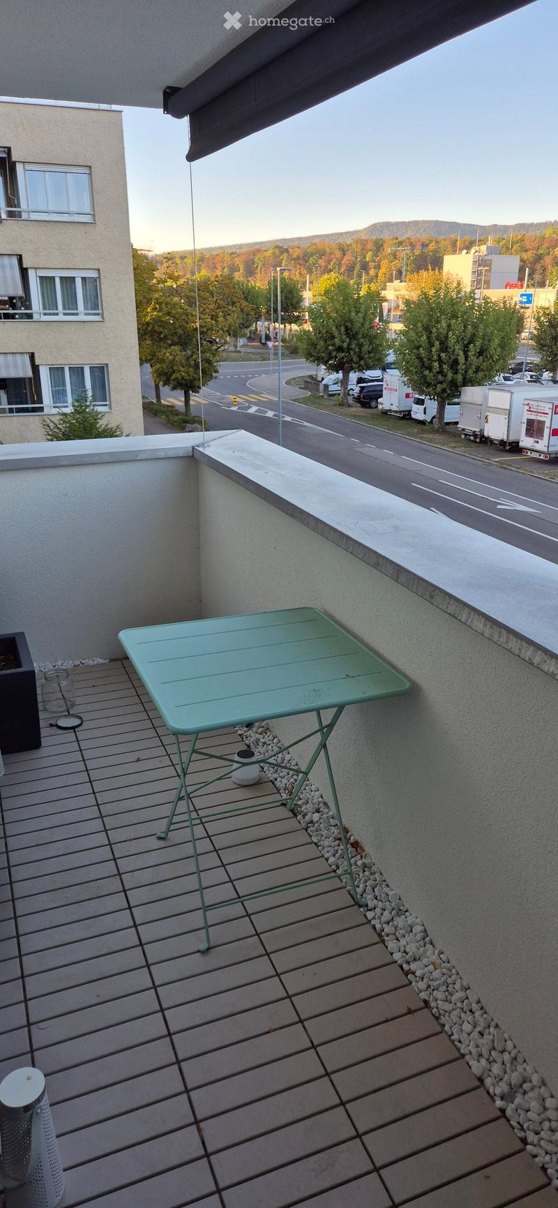 Balcony with wooden decking, green metal table, small potted plant, and a view of the street and surrounding area.