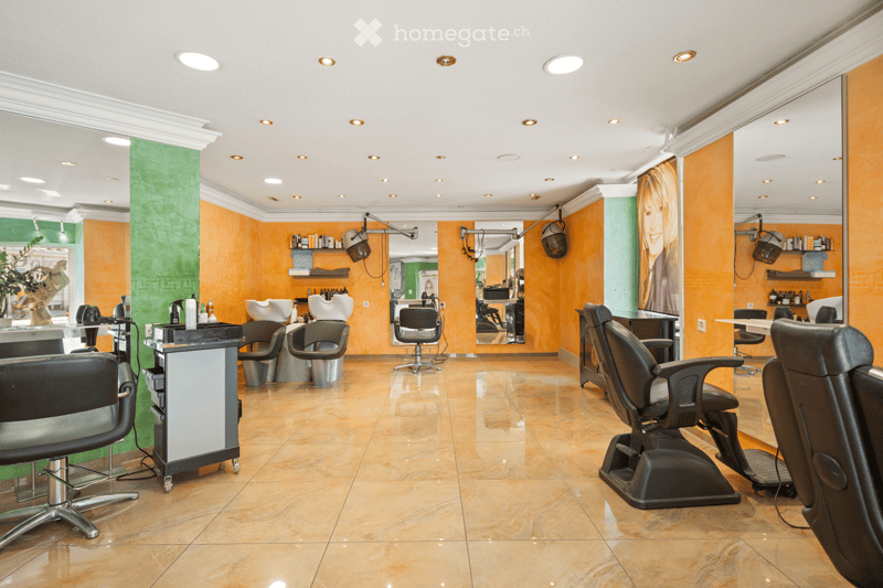 Modern salon with orange and green walls, multiple black salon chairs, mirrors, shelves with hair products, and a reception desk.