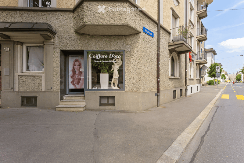 A multi-story building with a textured facade, featuring a storefront with a large advertisement for 'Coiffure D'oro' and a small entrance with steps.