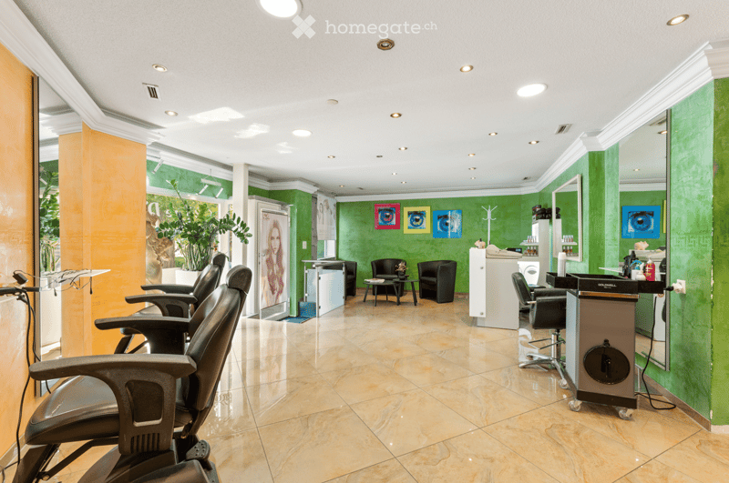 Modern salon with green walls, multiple black salon chairs, a reception desk, small tables, and a potted plant near the window.