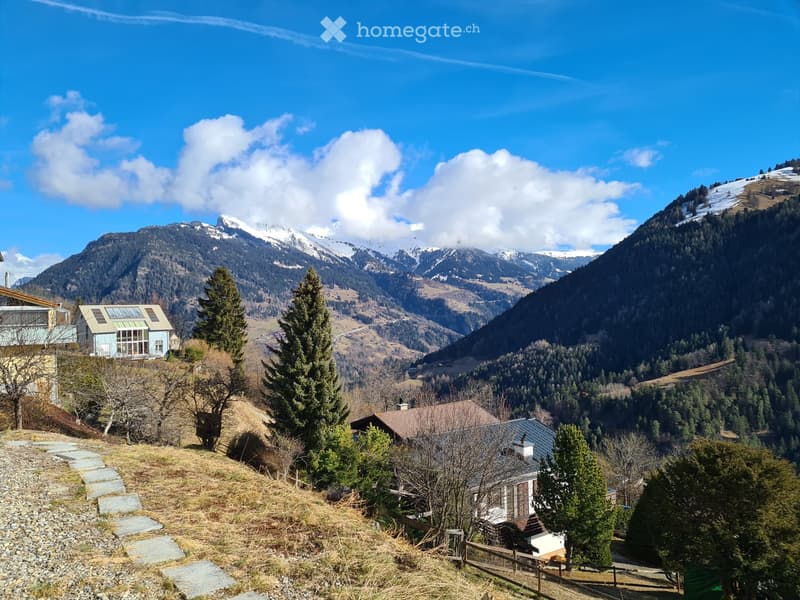 Scenic view of a house nestled in a mountainous landscape with snow-capped peaks in the background, surrounded by trees and a stone pathway.