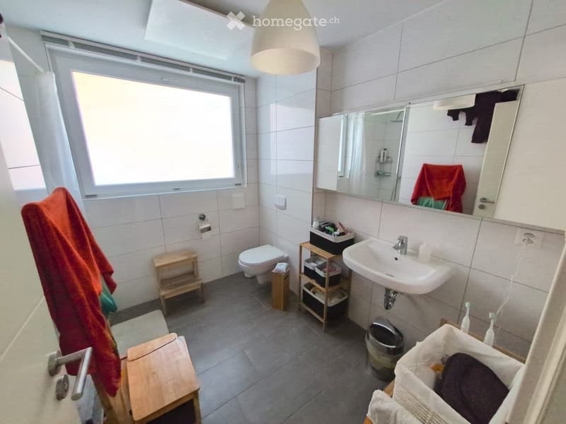 Modern bathroom with white tiles, a sink, toilet, mirror, and wooden stools. Features a large window, a hanging lamp, and a storage unit with baskets.