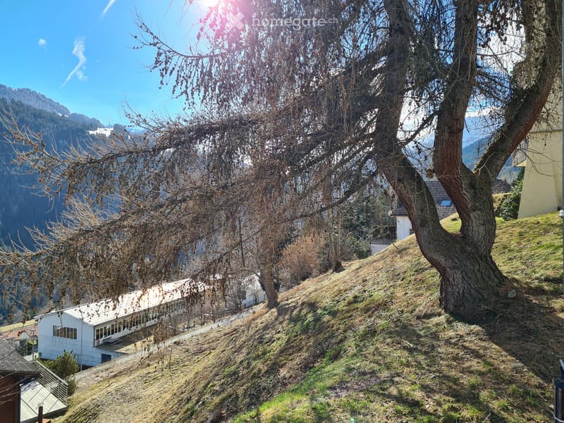 A scenic outdoor view with a modern building on a hillside, surrounded by trees and a mountainous backdrop under a clear blue sky.