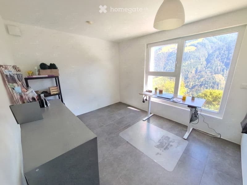 Bright room with large windows offering a scenic mountain view, featuring a sofa, a small table with a laptop, a shelf with books and decorative items, and a picture frame on the wall.