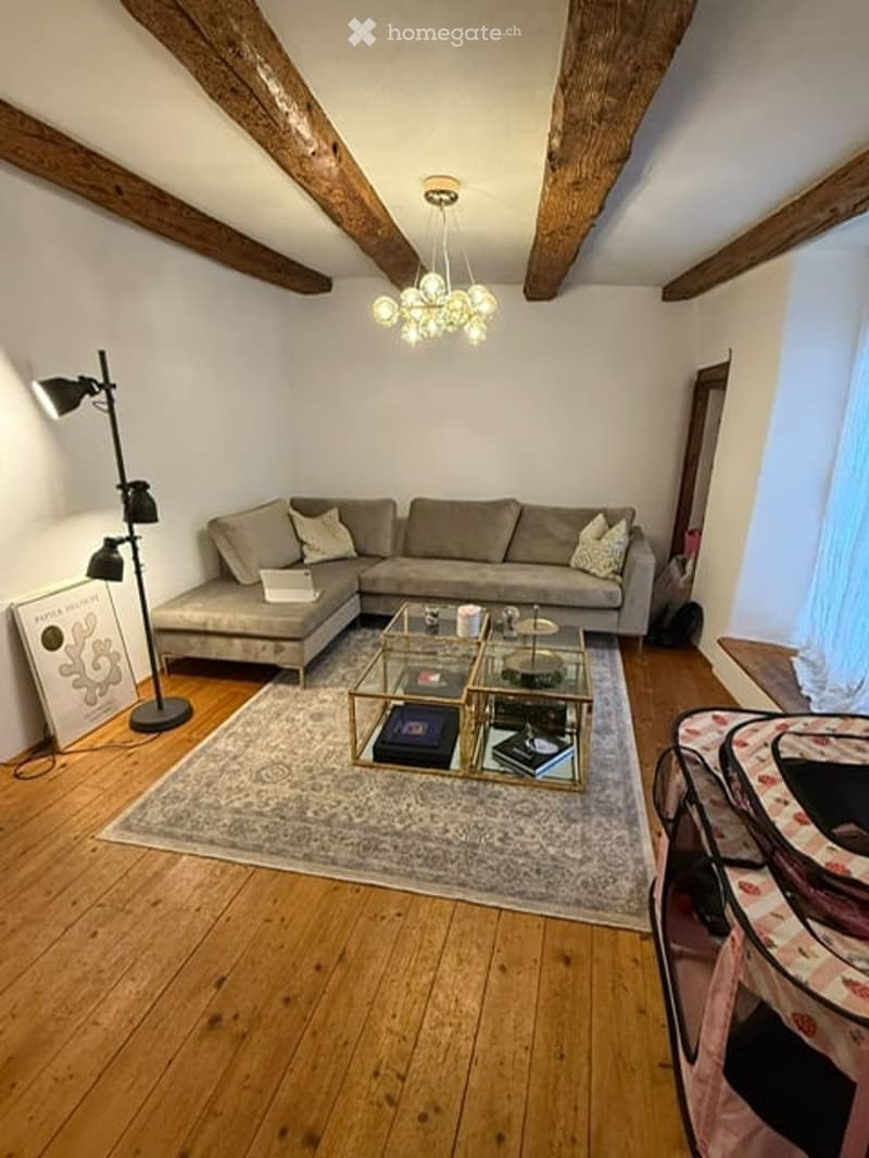 Living room with exposed wooden beams, grey sectional sofa, glass coffee table, floor lamp, and chandelier.