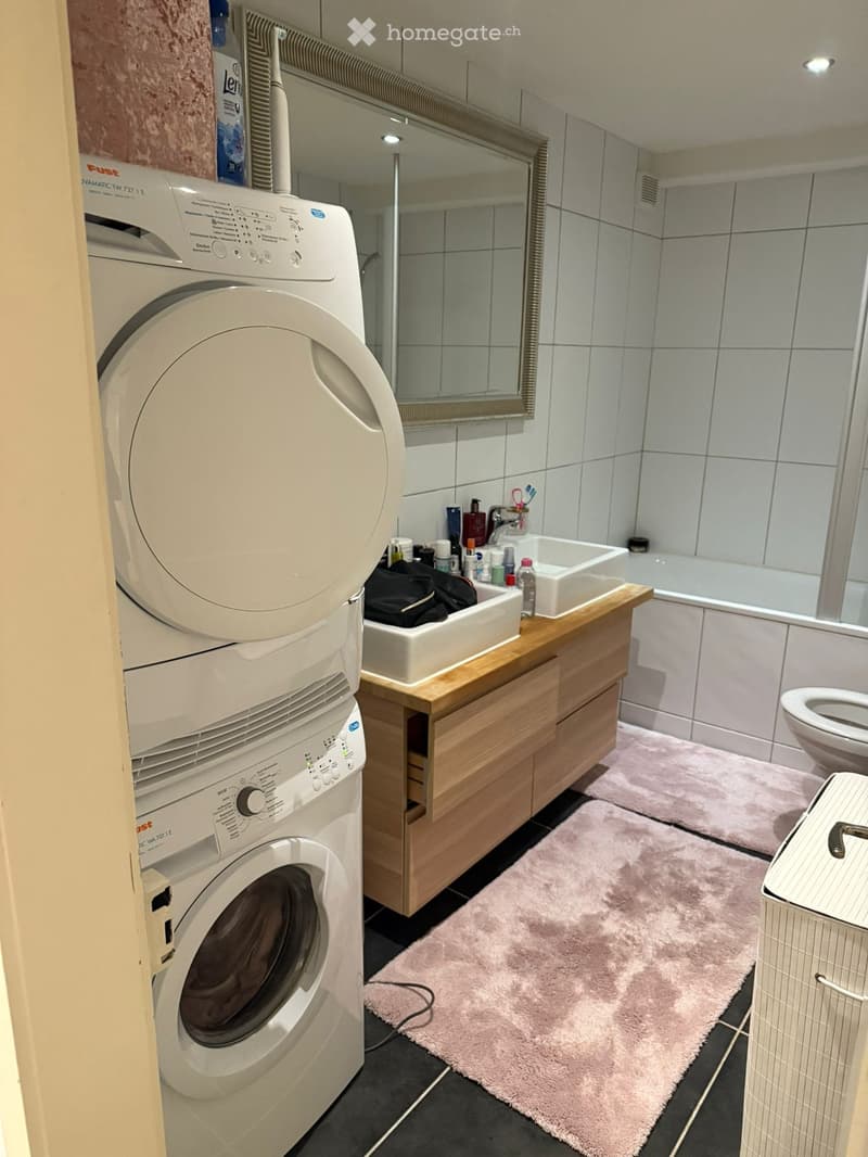 Modern bathroom with a washing machine, sink, toilet, bathtub, and a wooden cabinet. The floor is tiled, and there is a pink bath mat in front of the sink.