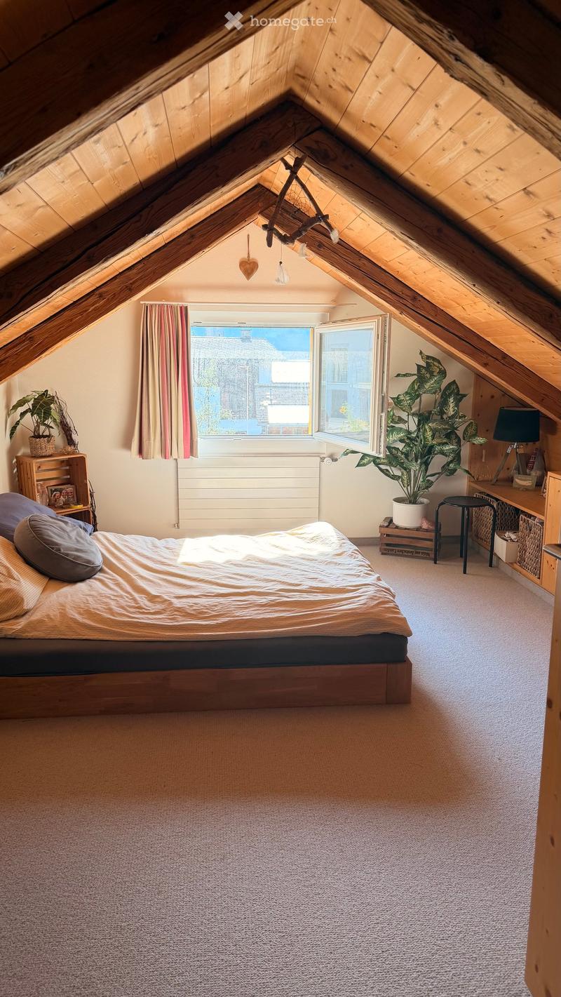 Cozy attic bedroom with wooden slanted ceilings, large windows with curtains, a bed with pillows and blankets, a nightstand with a lamp, a small stool, and potted plants.
