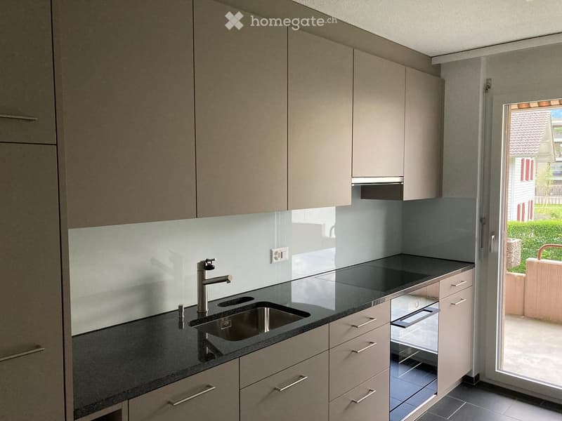 Modern kitchen with light-colored cabinets, a black countertop, a stainless steel sink, and a faucet. The space features a built-in oven and a sliding glass door leading to an outdoor area.
