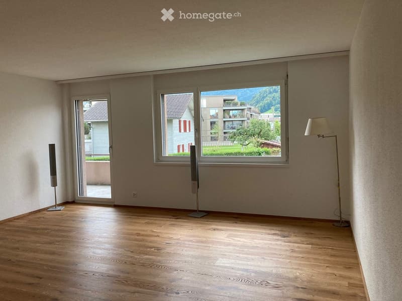 Empty room with wooden flooring, large windows, and a sliding door leading to a balcony. Two tall speakers are placed on the floor near the windows.
