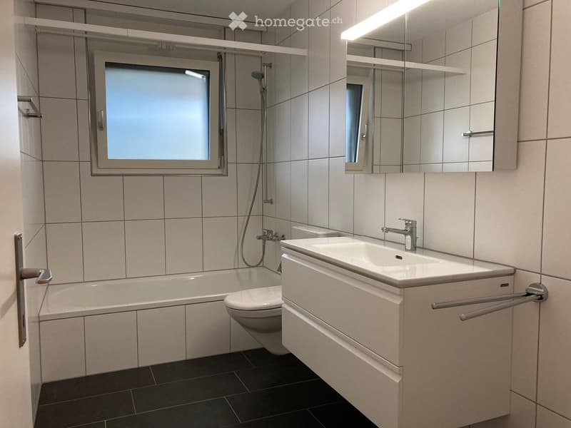 Modern bathroom with a bathtub, toilet, sink, and large mirror. Features tiled walls and a frosted window.