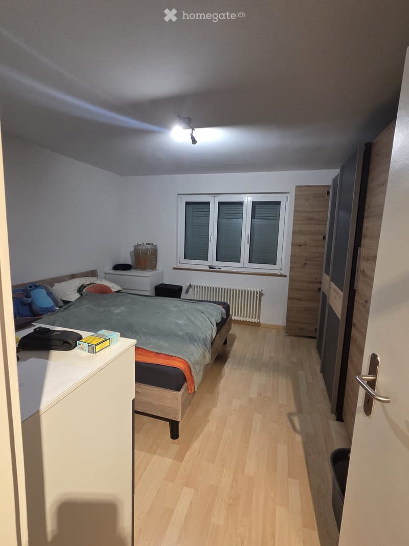 Cozy bedroom with wooden flooring, a double bed with pillows and blankets, a nightstand with a lamp, a window with blinds, and a radiator below it.