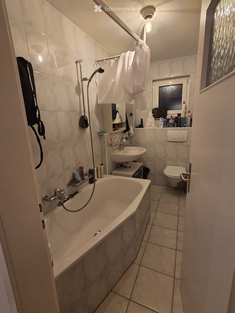 Bathroom with a bathtub, toilet, sink, and showerhead. Tiles on walls and floor, towel rack with towels, and a small window with frosted glass.