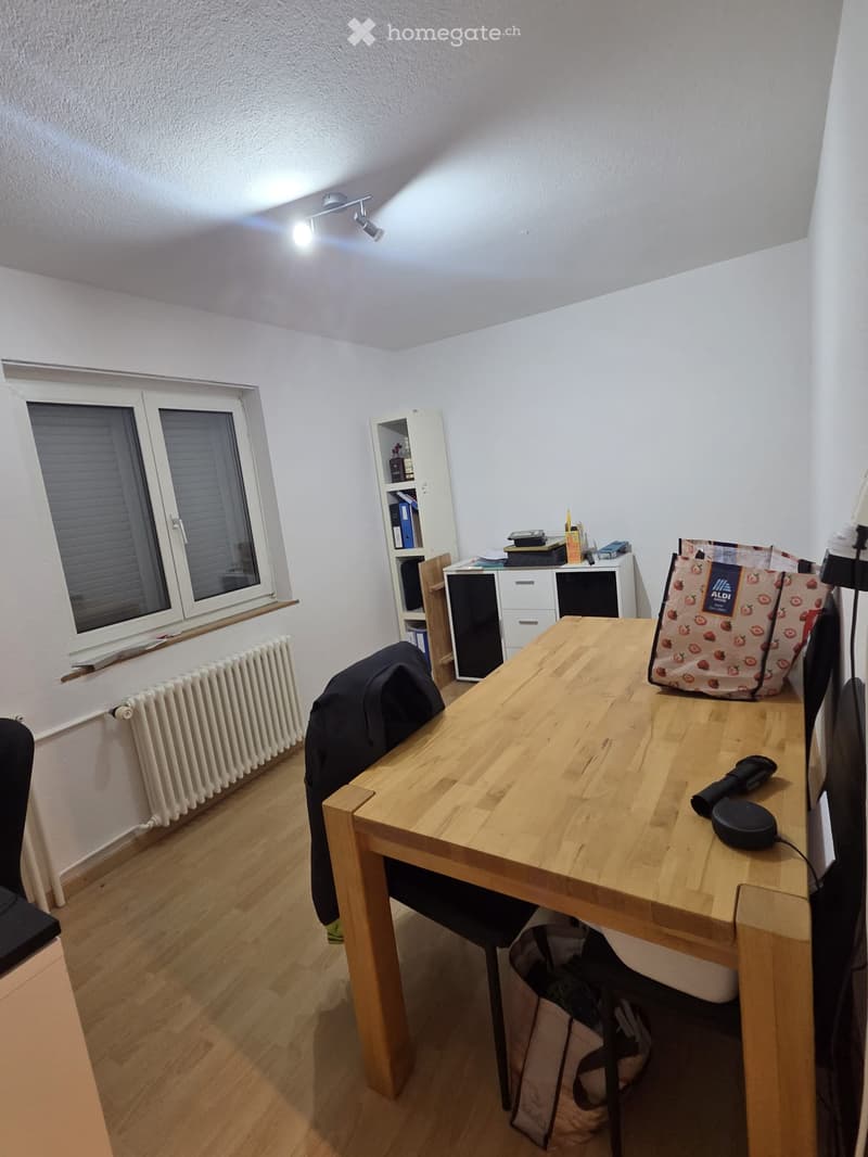 A well-lit room with wooden flooring, a large window with blinds, a wooden table with a laptop, a chair, a backpack, and a shelf with books and storage units.