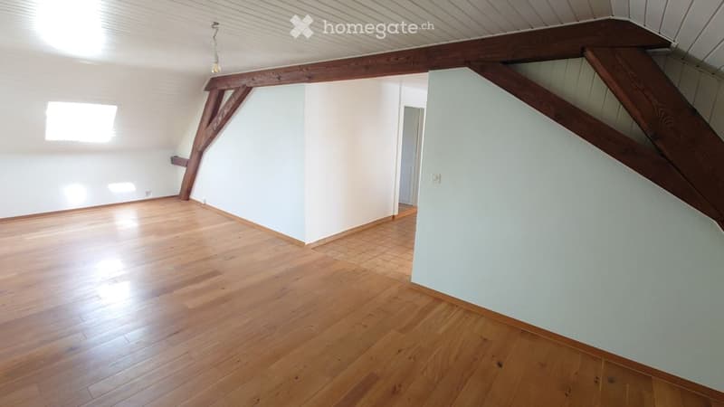 Empty attic room with wooden flooring, white walls, and exposed wooden beams. Natural light from a small window illuminates the space.