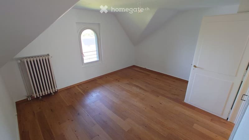 Empty attic room with wooden flooring, white walls, a single window, and a radiator. No furniture or equipment present.