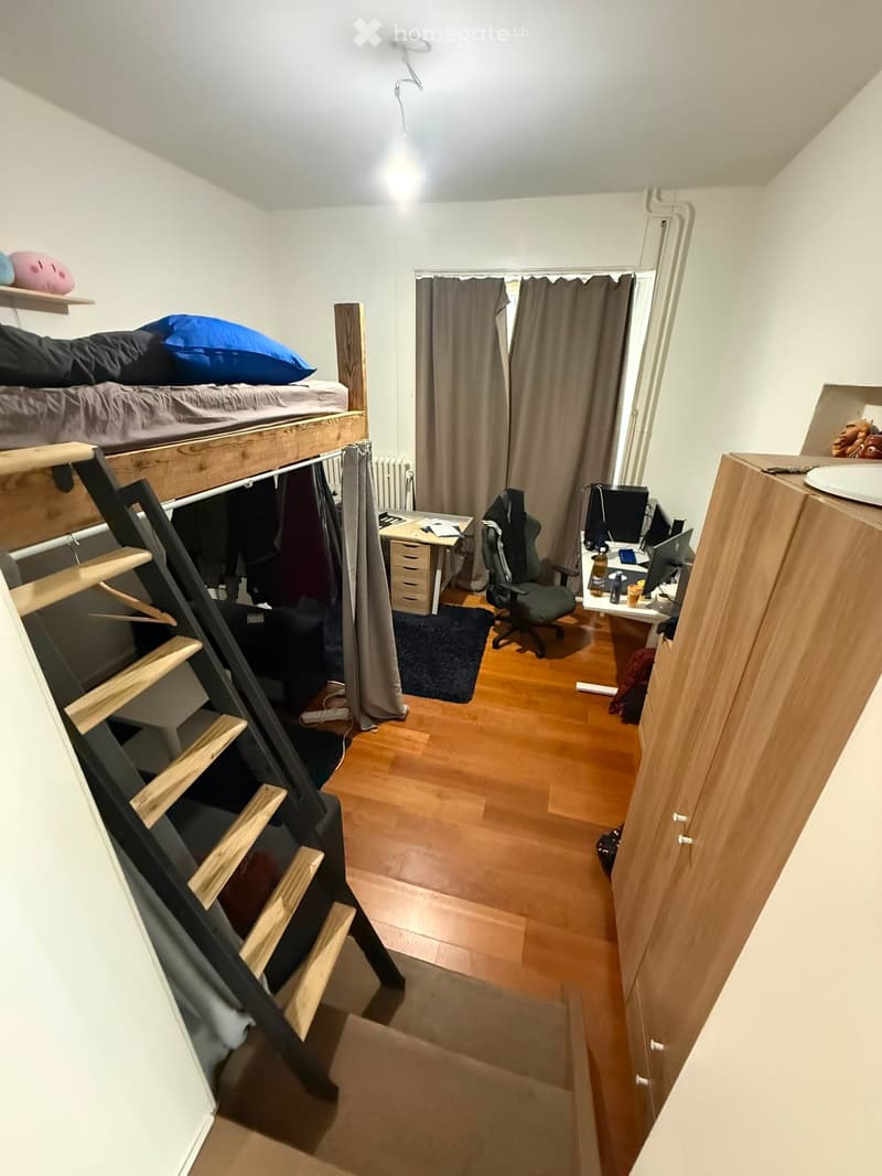 A compact, furnished room with a loft bed, desk, chair, and laptop. The room features wooden flooring, a curtained window, and shelves with decorative items.