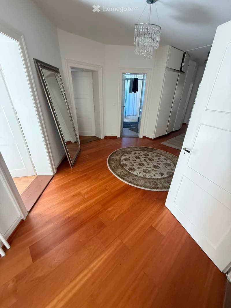 Empty hallway with wooden flooring, white walls, a large mirror on the left, a round patterned rug in the center, and several doors leading to other rooms.