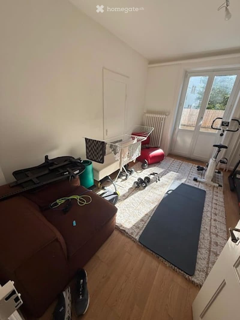 A multi-purpose room with a brown sofa, exercise bike, yoga mat, radiator, laundry rack, dumbbells, guitar, and a small stool. The room has wooden flooring and large windows letting in natural light.