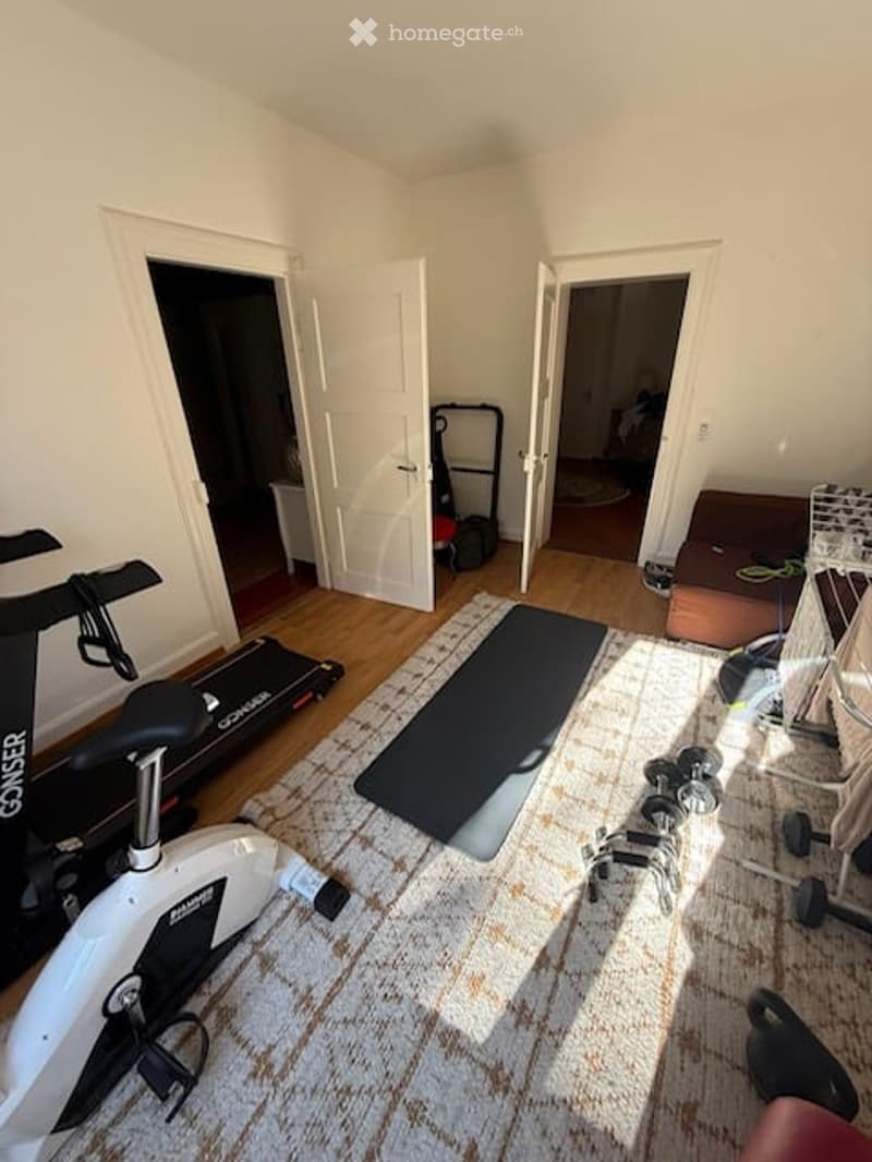 Well-lit room with wooden flooring, exercise bike, treadmill, yoga mat, dumbbells, and storage rack.
