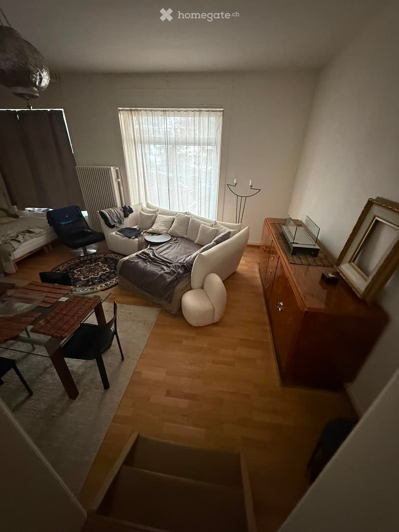A modern living room with a white sectional sofa, a wooden dining table, a black chair, a floor lamp, and a mirror on a wooden cabinet.