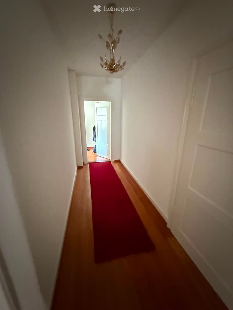 A narrow hallway with wooden flooring, white walls, and a red runner. A chandelier hangs from the ceiling, and an open door at the end leads to another room.