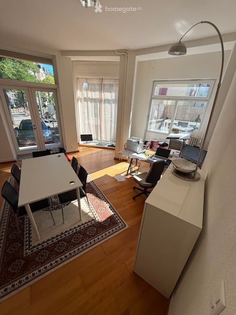 Bright, open-plan room with wooden flooring, large windows, dining table with chairs, work area with laptops, and a tall floor lamp.
