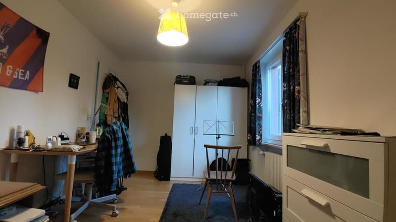 A furnished room with a wooden floor, a large window with curtains, a white cabinet, a desk with a laptop, a chair, and a lamp hanging from the ceiling.