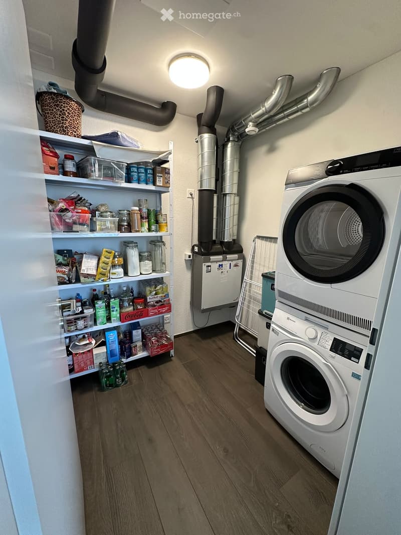 A small utility room with wooden flooring, white walls, and a ceiling light. Contains a washing machine, a drying machine, shelves filled with various items, and storage bins.