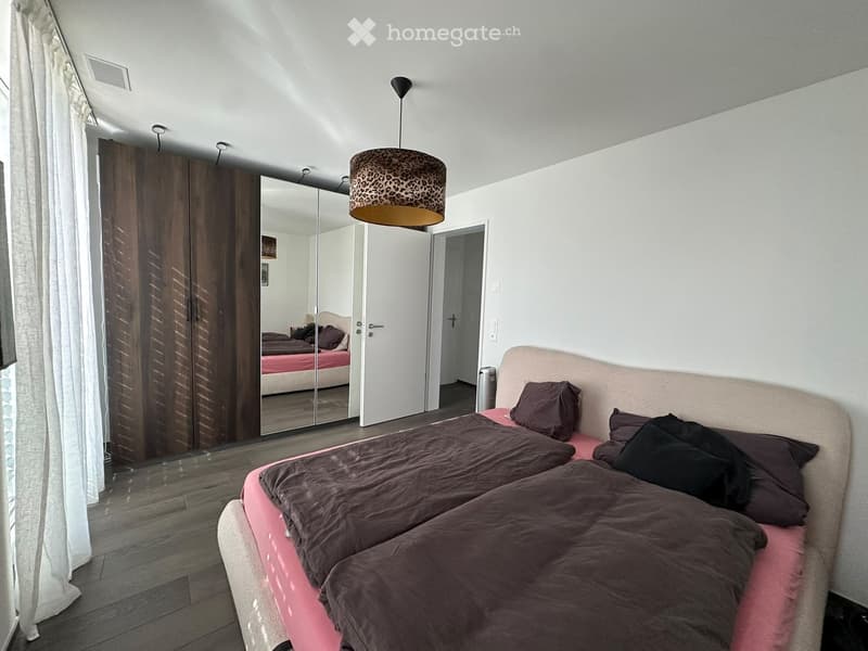 Modern bedroom with a large bed, dark bedding, a wardrobe with mirrored doors, and a hanging lamp with a leopard-print shade.