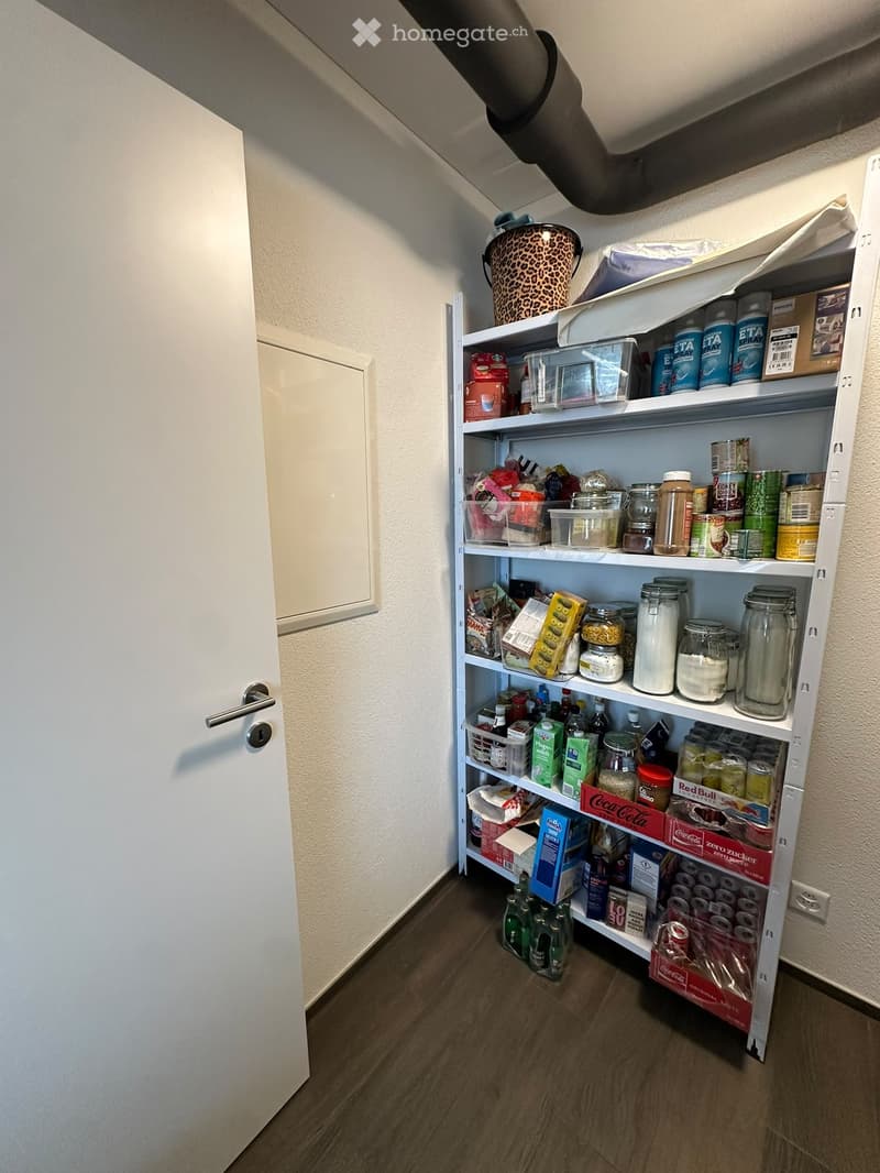 A pantry or storage room with white shelves filled with various food items, drinks, and containers. The room has a wooden floor and white walls.