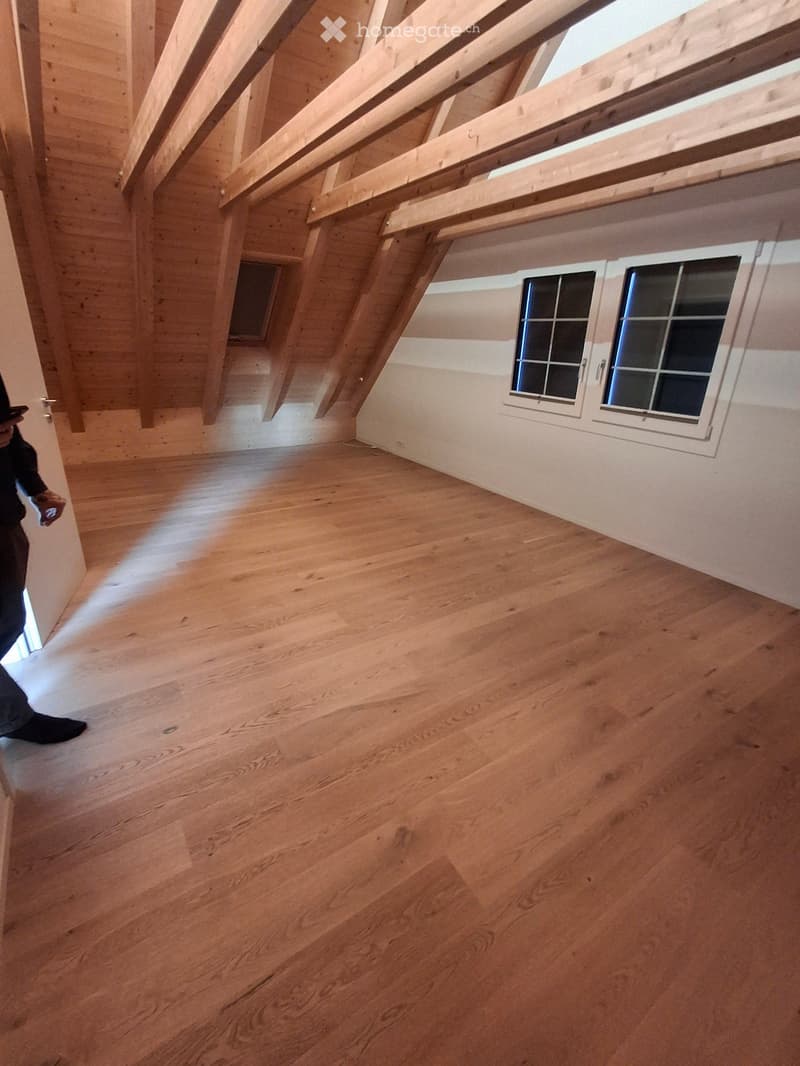 Empty attic space with wooden floors and exposed wooden beams, featuring two windows on one wall and a doorway leading to another area.