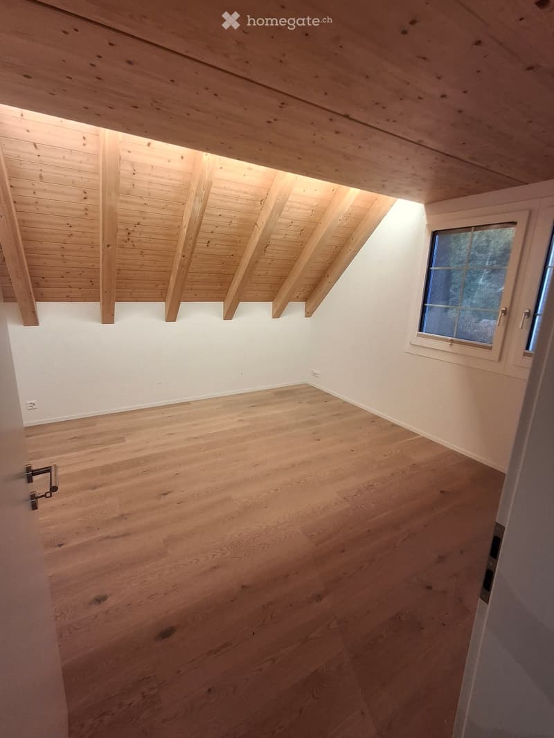 Empty room with wooden floor and exposed wooden ceiling beams, featuring a single window with a view of trees.