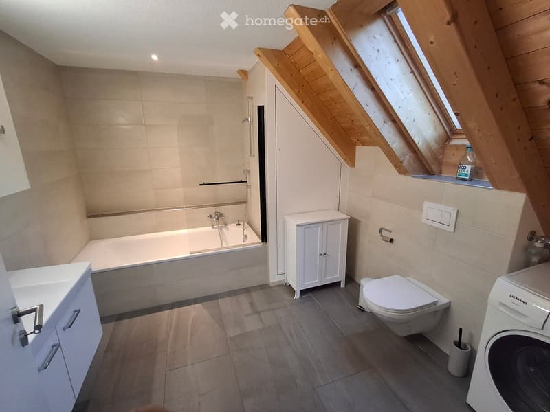 Modern bathroom with a bathtub, toilet, sink, cabinet, and washing machine. Features wooden slanted ceilings and large windows.
