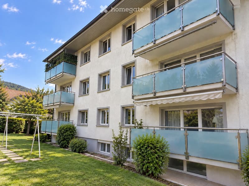 Multi-story residential building with light-colored facade, multiple windows, and glass-enclosed balconies. Green lawn and shrubs in front.