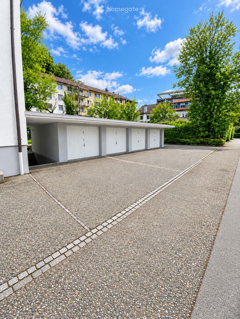 A modern, covered garage with four white doors, situated in a residential area with trees and neighboring buildings visible in the background.