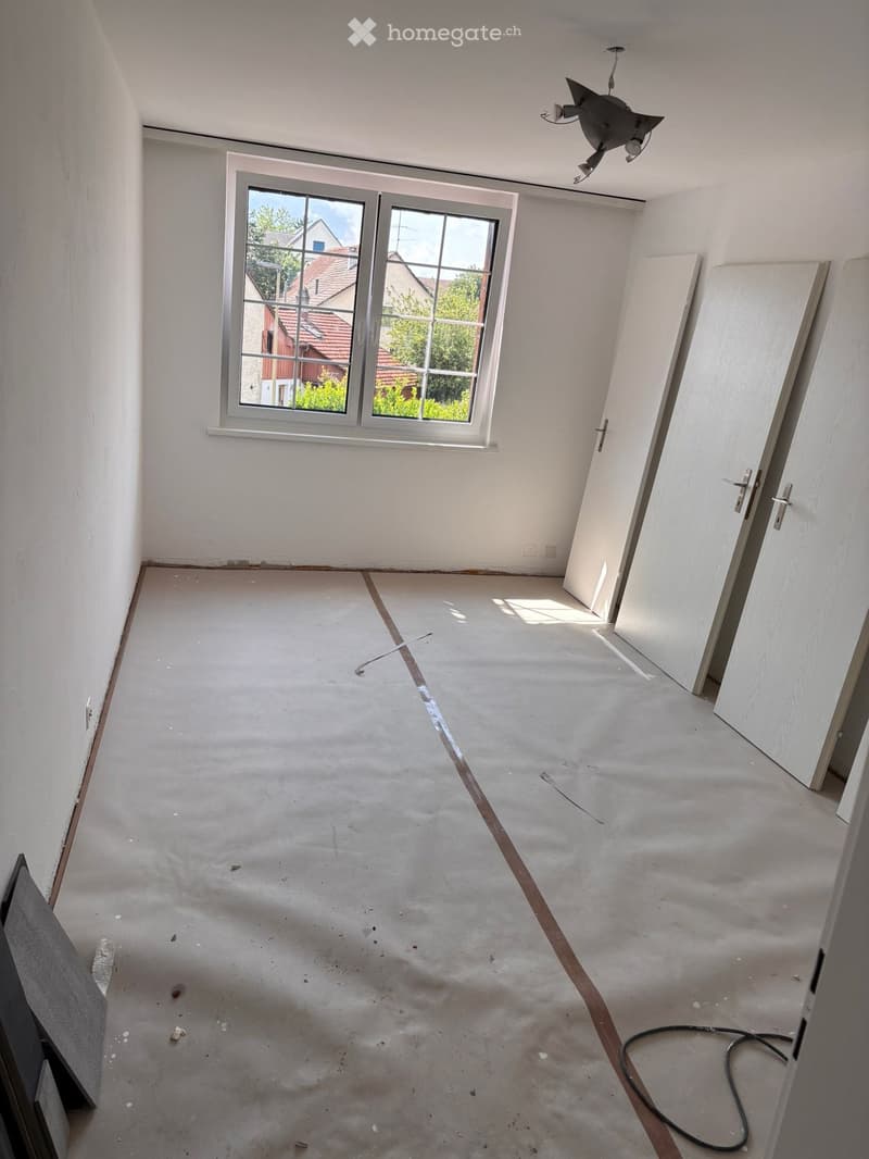 Empty room with bare walls and concrete floor, featuring a large window and several doors.