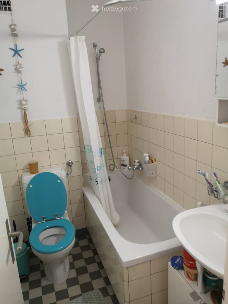 Bathroom with a white bathtub, a toilet with a turquoise seat cover, a white sink, and a toothbrush holder. The walls are tiled in beige and white, and the floor has a black and white checkered pattern.