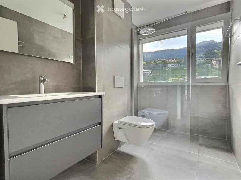 Modern bathroom with a sink, faucet, mirror, cabinet, and toilet, featuring large windows with a scenic mountain view.