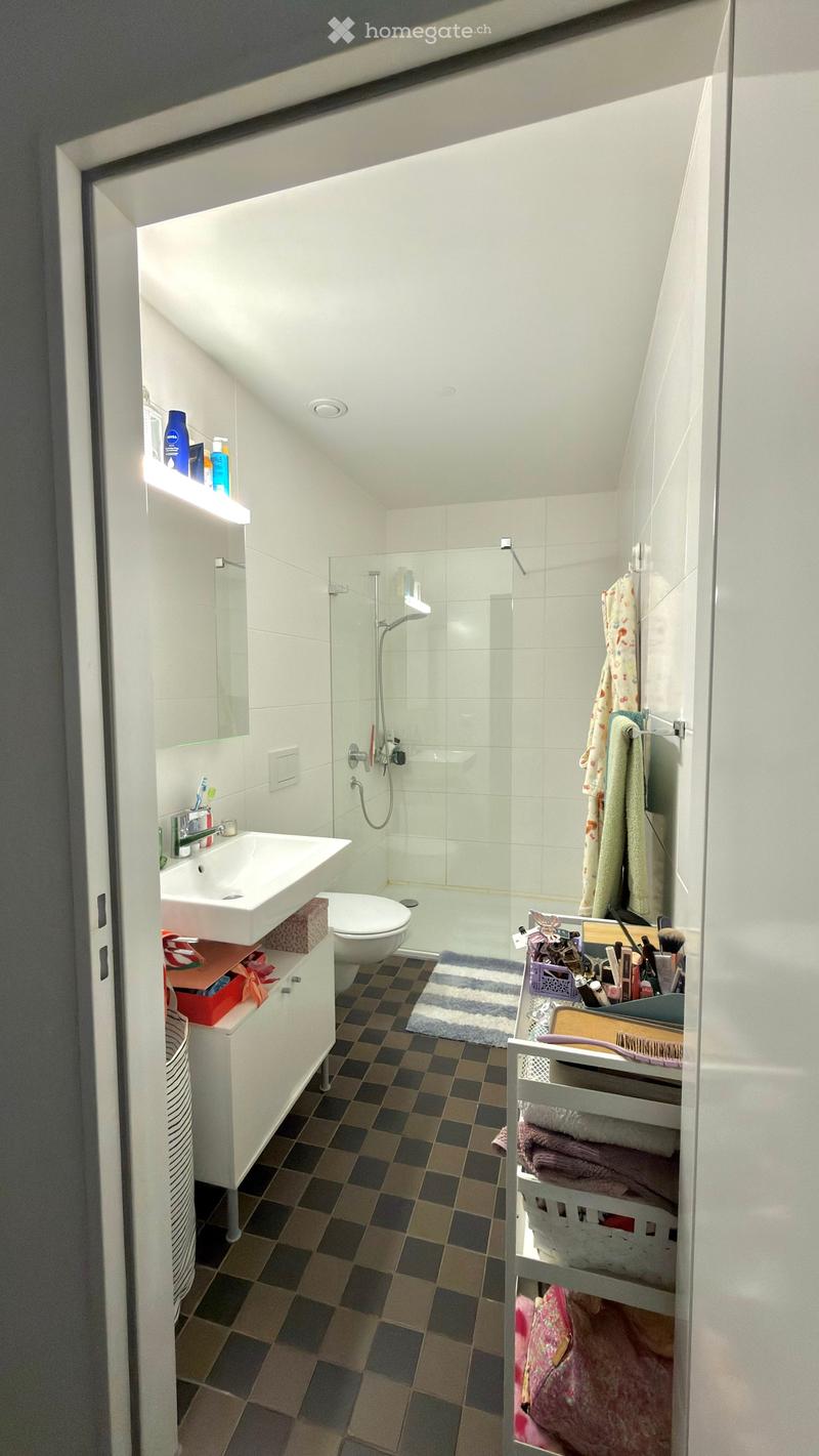 Modern bathroom with white tiles, a glass-enclosed shower, a sink, a toilet, and various personal care items on shelves and a basket.
