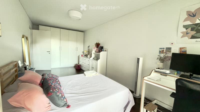 A modern bedroom with a neatly made bed, a desk with a computer, a wardrobe, and decorative items on shelves.